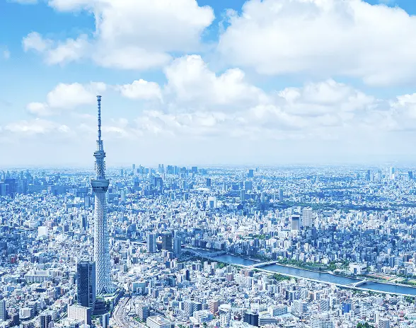 青空の下、東京スカイツリーとその周辺の都市景観を捉えた空撮写真。広がる高層ビル群と川の流れが都市の広大さを表現している。