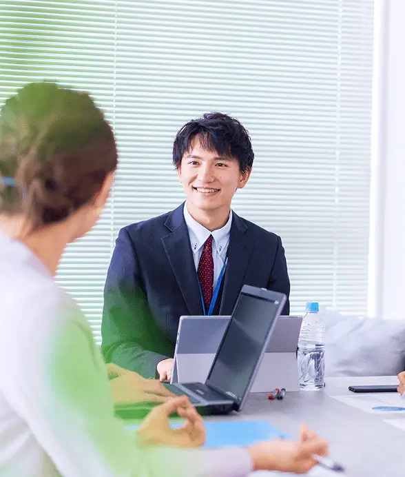 スーツ姿の笑顔の男性が、パソコンを使いながら女性と話している様子。