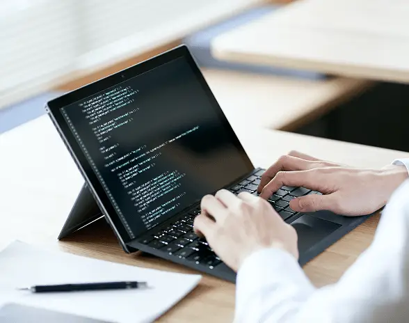 テーブルの上に置かれたノートパソコンでプログラミングコードを入力している手元のアップ。白いシャツの袖と木目のデスクが映る仕事や開発の一場面。