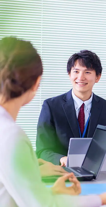 スーツ姿の笑顔の男性が、パソコンを使いながら女性と話している様子。